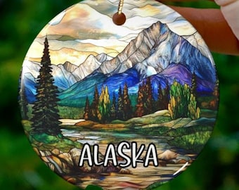 May include: A round ceramic ornament with a stained glass style image of a mountain range and a river. The ornament is decorated with a blue, green, and purple color scheme. The text "ALASKA" is printed in white on the ornament.