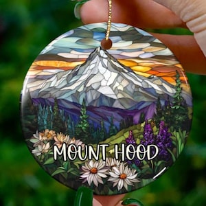 May include: A round ceramic ornament with a stained glass design of Mount Hood, a mountain in Oregon. The ornament features a colorful sunset sky, a snow-capped mountain, and a green forest with wildflowers. The text "MOUNT HOOD" is printed in black on the ornament.