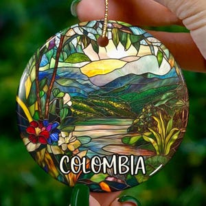 May include: A round ceramic ornament with a stained glass design depicting a landscape with mountains, a river, and flowers. The ornament is decorated with the word "COLOMBIA" in a stylized font.