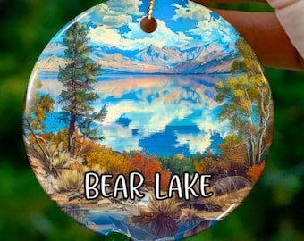 Puede incluir: Adorno navideño redondo con un paisaje del lago Bear. El adorno muestra una escena vibrante del lago con montañas, árboles y el texto "Bear Lake". El adorno es sostenido por una mano con esmalte de uñas verde.