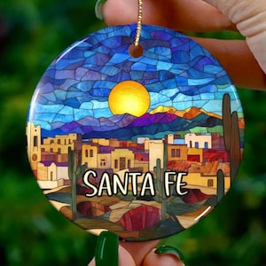 Könnte beinhalten: Runder Keramikschmuck mit einem Design im Glasmalerei-Stil von Santa Fe, New Mexico. Das Design umfasst eine gelbe Sonne, blauen Himmel, Berge, Gebäude und Kakteen. Die Worte "SANTA FE" sind in Weiß gedruckt.