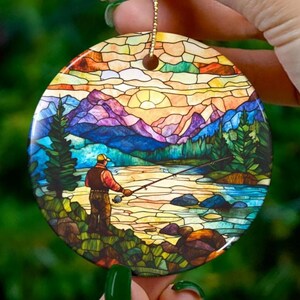 May include: A round, stained-glass style Christmas ornament featuring a fisherman in a mountain landscape. The ornament has a gold string for hanging. The design includes a river, trees, mountains, and a colorful sky.