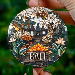 May include: Round ceramic ornament featuring a colorful Bali temple scene with the word "BALI" in a bold font. The design includes flowers, trees, and a temple, with a gold string for hanging. The ornament is held by a hand.
