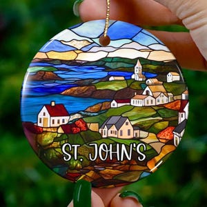 Op de afbeelding: Ronde keramische ornament met een glas-in-lood ontwerp van een kustplaats met kleurrijke gebouwen en een blauwe watermassa. De ornament heeft de tekst "ST. JOHN'S" erop gedrukt.