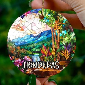 May include: A round ceramic ornament with a stained glass design of a tropical landscape. The ornament features a waterfall, lush greenery, and palm trees. The text "HONDURAS" is printed on the bottom of the ornament.