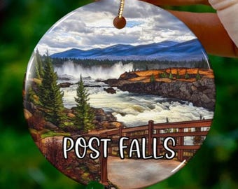 Puede incluir: Un adorno redondo de cerámica con una imagen escénica de una cascada en un entorno boscoso. El adorno presenta un puente de madera y el texto "POST FALLS".