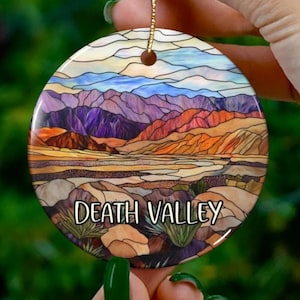May include: A round ceramic ornament with a stained glass design of a mountain range and desert landscape. The ornament has the text "DEATH VALLEY" printed on the bottom.
