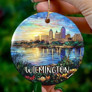 May include: A round ceramic ornament with a stained glass style image of the Wilmington skyline at sunset. The ornament has a gold string for hanging and the text "WILMINGTON" is printed on the bottom.