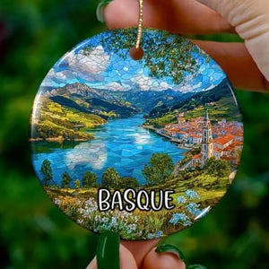 May include: Circular ceramic ornament depicting a landscape with a river, mountains, and a town. The ornament is blue, green, and white, with the word "BASQUE" in white text.