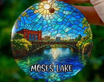 Könnte beinhalten: Runder Weihnachtsschmuck mit einem Buntglas-Stil, der Moses Lake, Washington, zeigt. Das Design umfasst eine Brücke, Gebäude und Wasser, mit dem Text "MOSES LAKE" unten. Der Schmuck hat eine goldene Aufhängeschnur.