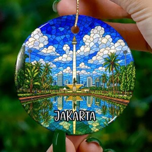 Peut inclure: Un ornement rond en céramique présentant une illustration de style vitrail du Monument National de Jakarta, en Indonésie. Le motif comprend un grand obélisque central, des palmiers, des bâtiments urbains et un bassin, avec le texte JAKARTA.