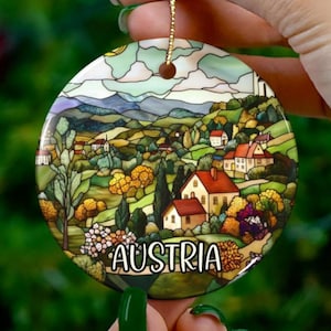 May include: A round ceramic bauble featuring a stained glass style image of an Austrian village. The ornament depicts a colourful landscape with houses, trees and mountains. The word "Austria" is written in white lettering at the bottom of the bauble.