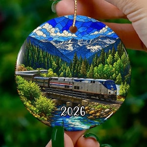 May include: A round ceramic ornament featuring a stained-glass style illustration of a passenger train crossing a bridge over a river. The scene includes a dense green forest, snow-capped mountains, a blue sky, and the year 2026 printed in black.