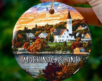 Könnte beinhalten: Ein runder Keramik-Anhänger mit einem Bild im Stil eines Buntglasfensters von einer kleinen Stadt mit einem Kirchturm. Der Anhänger hat eine goldene Schnur zum Aufhängen und den Text "Mackinac Island" unten aufgedruckt.