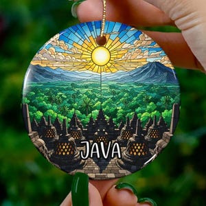 Op de afbeelding: Een rond keramisch ornament met een illustratie in glas-in-loodstijl van de Borobudur-tempel op Java, Indonesië. Het ontwerp toont een heldere gele zon boven een groen bos, bergen en stenen stoepa's, met het woord JAVA erop gedrukt.