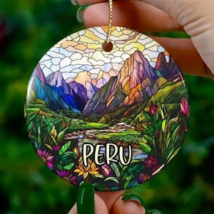 Puede incluir: Adorno navideño redondo de cerámica con un colorido paisaje de montañas, un río y exuberante vegetación. La palabra "PERU" está escrita en blanco. El adorno está sostenido por una mano con esmalte de uñas verde.