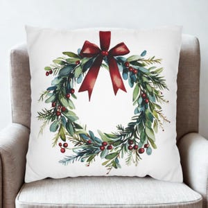 May include: A white cushion with a watercolour design of a Christmas wreath. The wreath is made of green leaves, red berries, and pine needles, and is tied with a large red bow. The cushion is on a neutral-toned armchair.