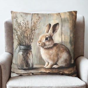 May include: A decorative square pillow featuring a painted image of a brown rabbit sitting next to a glass jar filled with dried plants. The background has a rustic, wooden panel design. The pillow is placed on a light-colored chair.
