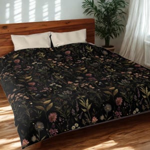 Peut inclure: Une housse de couette florale noire avec un motif botanique détaillé. Le motif comprend diverses fleurs et feuillages dans des tons de rose, de vert et de beige, sur un fond sombre. La literie est présentée sur un cadre de lit en bois.