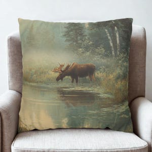 Puede incluir: Cojín cuadrado con un paisaje sereno con un alce de pie en un cuerpo de agua brumoso. La obra de arte representa un alce marrón con grandes astas, rodeado de árboles y hierba alta. La paleta de colores incluye verdes, marrones y azules.
