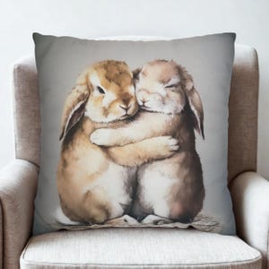 Könnte beinhalten: Ein quadratisches Zierkissen mit einer Aquarellillustration von zwei sich umarmenden Hasen. Die Hasen sind in Braun- und Beigetönen gehalten, mit weichem Fell und geschlossenen Augen, vor einem grauen Hintergrund. Das Kissen liegt auf einem neutralen Stuhl.