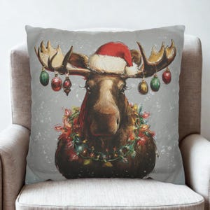 May include: A square throw pillow featuring a brown moose illustration on a light grey background. The moose wears a red Santa hat and has colourful Christmas ornaments hanging from its antlers, with a string of glowing lights around its neck.