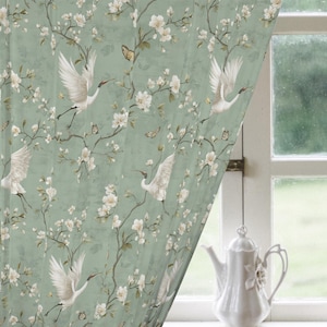 Puede incluir: Cortina verde salvia con un patrón repetido de grullas blancas, mariposas y ramas florecidas. La cortina cubre parcialmente una ventana, con una tetera blanca en el alféizar. El diseño tiene una estética vintage y botánica.