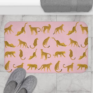 May include: Pink rectangular bath mat featuring a repeating pattern of stylized cheetahs in various poses. The cheetahs are tan with black spots. The mat is rectangular and has rounded corners. A pair of gray slippers are in the lower left corner.