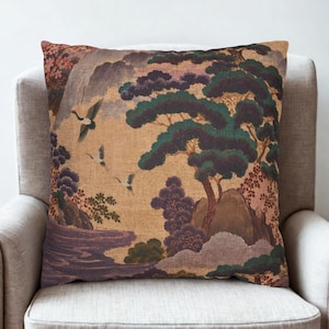 May include: A square throw pillow featuring a printed landscape scene with three flying cranes, dark green pine trees, purple rock formations, and a flowing river against a textured tan background. The design includes detailed foliage and mountains.
