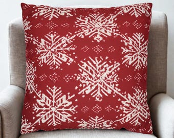 May include: A square cushion featuring a red backdrop adorned with a repeating design of white snowflakes. The cushion is positioned on a light grey armchair, creating a festive atmosphere. Ideal for adding a touch of winter charm to any living space.