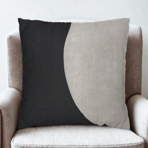 May include: Square decorative pillow with a minimalist design. The pillow features a black section on the left and a light gray, crescent-shaped section on the right. The pillow is placed on a beige armchair.