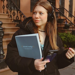 Può includere: Una persona con cuffie nere e una giacca nera tiene in mano un taccuino con la scritta "dream nudge" e uno smartphone. Lo sfondo presenta un edificio in pietra marrone con scale e una ringhiera in ferro battuto.