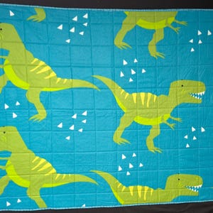 May include: A turquoise quilt featuring a repeating pattern of green and yellow dinosaurs. The dinosaurs have sharp teeth and are in various poses. White triangle shapes are scattered across the quilt.