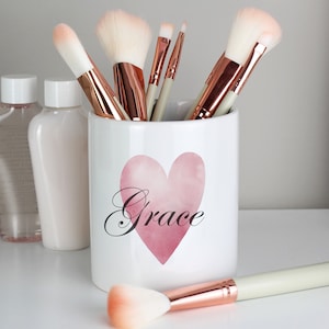Puede incluir: Portacepillos de maquillaje de cerámica blanca con un corazón rosa acuarela y la palabra "Grace" en escritura negra. El portacepillos está lleno de varios pinceles de maquillaje con mangos de oro rosa.