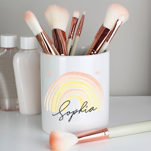 May include: White ceramic makeup brush holder with a rainbow design and the name "Sophia" written in black script. The holder is filled with various makeup brushes.