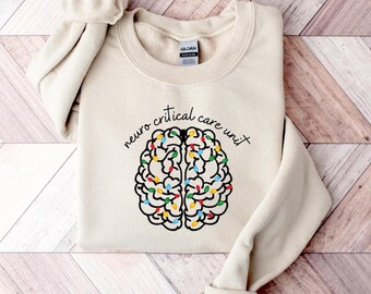 May include: A cream-colored sweatshirt with the words "neuro critical care unit" printed above a brain graphic. The brain illustration is outlined in black and filled with colorful lights, creating a festive and medical theme.