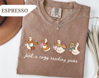 May include: A brown short-sleeved t-shirt with a graphic print of four white geese wearing scarves and carrying books. The text "just a cozy reading goose" is printed below the geese. The shirt has a crew neckline and a Comfort Colors brand label.