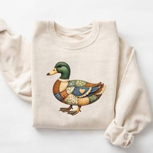 May include: A cream-colored sweatshirt with a colorful patchwork duck design. The duck features a green head, yellow beak, and a body composed of various fabric patterns in shades of brown, blue, and green. The sweatshirt is folded.