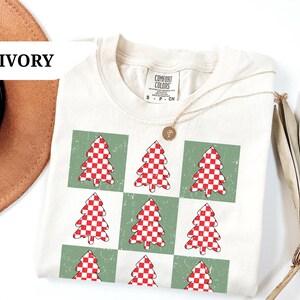 May include: Ivory-colored t-shirt featuring a grid pattern of green squares, each containing a red and white checkered Christmas tree design. The shirt has a crew neck and short sleeves. A gold necklace with a pendant is draped on the shirt.