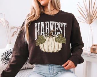 May include: A dark brown long-sleeve sweatshirt featuring a graphic print of a white pumpkin with green leaves and the word HARVEST in a distressed white font centered above the pumpkin. The garment has a crew neckline and ribbed cuffs.