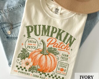 May include: An ivory t-shirt featuring a graphic print with the text PUMPKIN Patch, FRESH FROM THE PATCH, FARM FRESH, LOCALLY GROWN, and PUMPKINS CORN MAZE APPLE CIDER. The design includes a large orange pumpkin, green leaves, and a small flower.