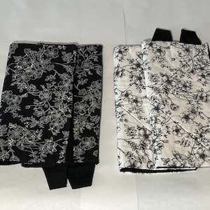May include: Two quilted fabric items, one black with white floral pattern, the other white with black floral pattern. Both have black accents and appear to be padded. The floral design features detailed blossoms and foliage.