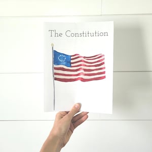 Puede incluir: Una tarjeta blanca con el texto "The Constitution" impreso en negro. Una pintura de acuarela de la bandera estadounidense con 13 rayas y un círculo de estrellas en el cantón azul está en la tarjeta.