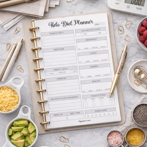 May include: A Keto Diet Planner notebook with gold spiral binding is open on a white surface. The planner includes sections for daily tracking of food intake, exercise, and sleep. Measuring cups with avocado and cheese are nearby, along with a digital scale and a bowl of raspberries.
