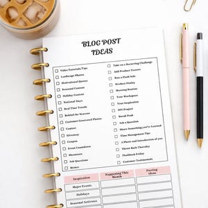 May include: A notebook with gold spiral binding, filled with blog post ideas, sits on a white surface. The notebook includes checkboxes and sections for inspiration and trends. A glass of iced coffee, gold paper clips, and pens are also present.