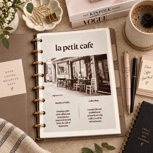 Op de afbeelding: Een planner met schijfbinding open op een pagina met de titel la petit cafe, met een zwart-witfoto van een café-exterieur. De pagina bevat tekst over de voordelen van koffie en een definitie van een koffieshop, naast een lichtroze en zwarte pen.