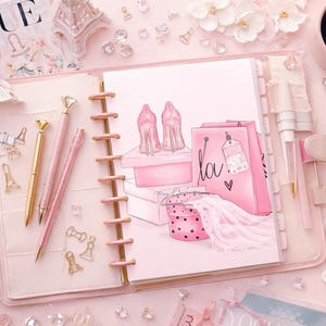 Puede incluir: Un planificador rosa con una ilustración de moda, bolígrafos y accesorios decorativos. La ilustración representa tacones altos, bolsas de compras y un vestido. También hay una revista Vogue, una figura de la Torre Eiffel y una taza de café.