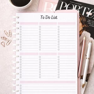 May include: A pink To Do List notebook with a white spiral binding, featuring a list format with checkboxes. A cup of coffee, pens, and a magazine are also visible, suggesting a workspace or desk setting.