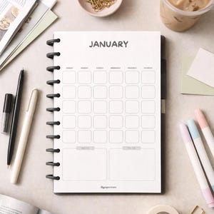 May include: A spiral-bound planner open to the January calendar page. The page has a grid layout for daily entries, with sections for notes and to-do lists. The image also includes pens, markers, and a cup of iced coffee.
