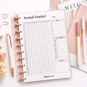 May include: A pink and white period tracker notebook with a grid layout, gold pen, and decorative paperclips. The notebook is open to a page labeled "Period Tracker". The image also includes pens and dried flowers.
